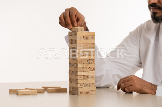 المتعه والتسلية وقضاء الوقت الممتع, ممارسة الأنشطة والألعاب المسلية, بورتريه مقرب ليد رجل عربي خليجي اماراتي يرتدي الكندورة يلعب الجينجا, محاولة رص الألواح الخشبية, تنمية مهارات التفكير والذكاء، خلفية بيضاء