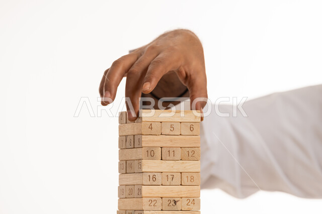 Developing thinking and intelligence skills, fun and entertainment, spending a good time, practicing entertaining activities and games, close-up portrait of the hand of an Arab Gulf Emirati man wearing a kandura playing Jenga, trying to stack wooden boards, white background