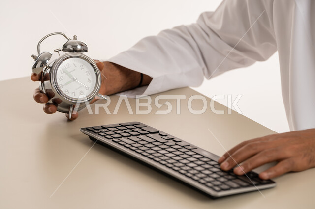 ادراك قيمة الوقت وانجاز المهام في الزمن المحدد, بورتريه مقرب لرجل عربي خليجي اماراتي يرتدي الكندورة يمسك بيده ساعة رقمية صغيرة, استخدام لوحة المفاتيح للكتابة, الدقة في المواعيد, مفهوم التذكير والتنبيه, خلفية بيضاء