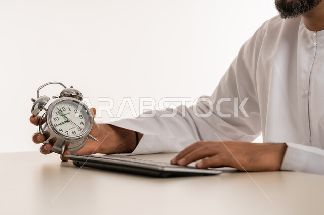 Using the keyboard for typing, punctuality, the concept of reminders and alerts, realizing the value of time and completing tasks on time, a close-up portrait of an Arab Gulf Emirati man wearing a kandura holding a small digital watch in his hand, white background