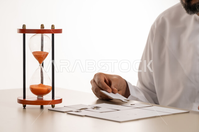 ألعاب الذكاء وتنمية المهارات, الدقة والتركيز وحل اللغز, بورتريه مقرب ليد رجل عربي خليجي اماراتي يرتدي الكندورة يحاول ربط وتوصيل قطع البزل بجانب الساعة الرملية الخشبية (البنكام), لعبة تركيب الصور, مفهوم قياس الوقت, التسلية وقضاء الوقت الممتع، خلفية بيضاء