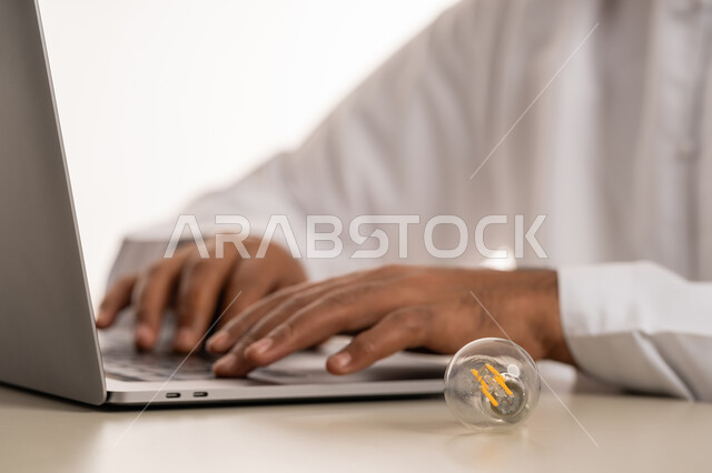 محاولة اكتشاف حلول للمشكلات, تطوير الأعمال والأفكار, محاولة تحقيق الانجازات, بورتريه مقرب ليد رجل عربي خليجي اماراتي يرتدي الكندورة يعمل على الحاسوب, استخدام الاجهزة والتقنيات الحديثة في انجاز المهام, مفهوم براءة الاختراع, المصباح الكهربائي الضوئي, خلفية بيضاء