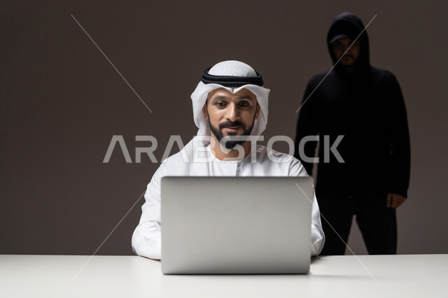 قرصنة وسرقة البيانات, التعرض للهجمات والاختراقات, التسلل عبر الأمن السيبراني، اختراق حماية وأمن وخصوصية المستخدمين, بورتريه لرجل عربي خليجي إماراتي يرتدي الكندورة والغترة البيضاء يستخدم جهاز الحاسوب المحمول ويقف ورائه المخترق, خلفية رمادية
