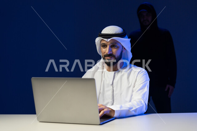 التعرض للهجمات والاختراقات, اختراق حماية وأمن وخصوصية المستخدمين, التسلل عبر الأمن السيبراني، قرصنة وسرقة البيانات, بورتريه لرجل عربي خليجي إماراتي يرتدي الكندورة والغترة البيضاء يستخدم جهاز الحاسوب المحمول ويقف ورائه المخترق, خلفية زرقاء
