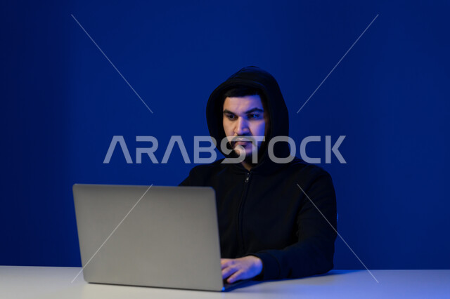 سرقة كلمات المرور والبيانات السرية، اختراق الأنظمة وحماية الأجهزة, التعرض للهجمات والاختراقات الالكترونية, مفهوم الجرائم الالكترونية, بورتريه مقرب لشاب عربي خليجي إماراتي يرتدي سترة سوداء يعمل في مجال الهكر الغير قانوني, خلفية زرقاء