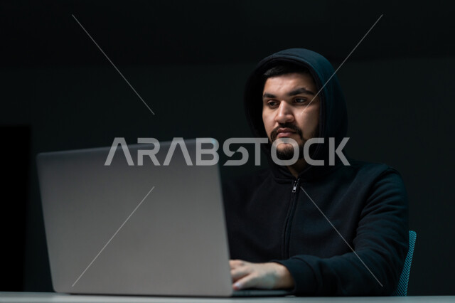 The concept of cybercrime, exposure to cyber attacks and hacking, hacking systems and protecting devices, stealing passwords and confidential data, close-up portrait of a young Emirati Gulf Arab man wearing a black jacket working in the field of illegal hacking, black background