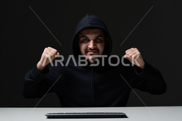 Cyber ​​security intrusion, data theft and hacking, computer used for illegal purposes, close-up portrait of young Arab Gulf Emirati man wearing black jacket working in hacking field raising his hands with victory expression, black background