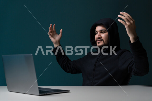 قرصنة وسرقة البيانات, التسلل عبر الأمن السيبراني, استخدم جهاز الحاسوب لأغراض غير قانونية, بورتريه مقرب لشاب عربي خليجي إماراتي يرتدي سترة سوداء يعمل في مجال الهكر يرفع يديه بتعابير الانتصار, خلفية زرقاء