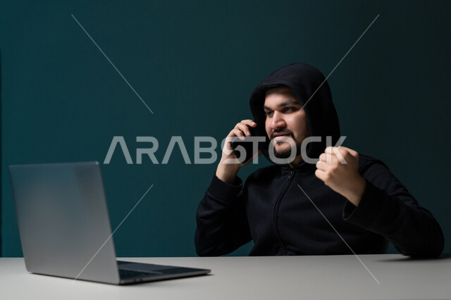 التجسس على المكالمات الهاتفية, استخدم جهاز الحاسوب لأغراض غير قانونية, التسلل عبر الأمن السيبراني، قرصنة وسرقة البيانات, بورتريه مقرب لشاب عربي خليجي إماراتي يرتدي سترة سوداء يعمل في مجال الهكر يرفع يده بتعابير الانتصار, خلفية زرقاء