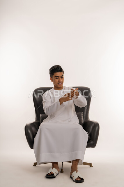 مفهوم الاناقة والرجولة والاهتمام بالمظهر الخارجي، بورتريه لشاب مراهق عربي خليجي اماراتي مبتسم يرتدي الكندورة يجلس على كرسي مريح يقوم بحركات يدين مختلفة بتعابير السرور، خلفية بيضاء
