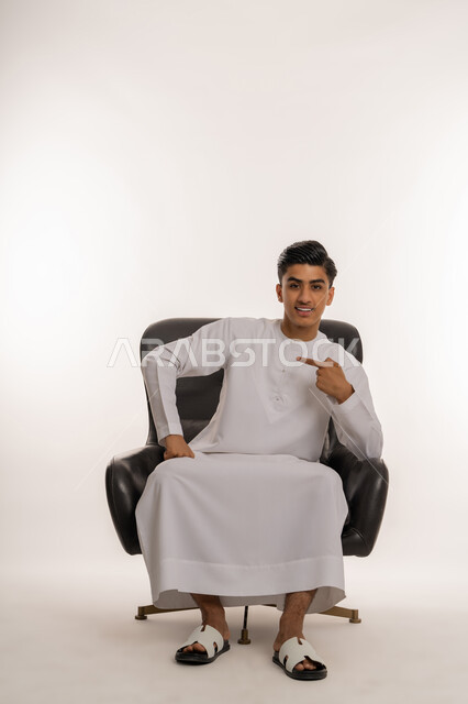 الاشارة بإصبع السبابة الى شيء ما، مفهوم الاناقة والرجولة والاهتمام بالمظهر الخارجي، بورتريه لشاب مراهق عربي خليجي اماراتي مبتسم يرتدي الكندورة يجلس على كرسي مريح ينظر الى الكاميرا بتعابير السرور والفرح، خلفية بيضاء