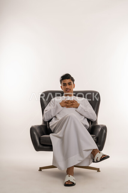 مفهوم الاناقة والرجولة والاهتمام بالمظهر الخارجي، تشابك الايدي مع بعضهم بتعابير التفكير، بورتريه لشاب مراهق عربي خليجي اماراتي مبتسم يرتدي الكندورة يجلس على كرسي مريح ينظر الى الكاميرا بتعابير السرور والفرح، خلفية بيضاء