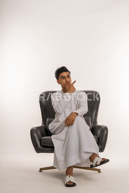 Feeling confused and uncertain, looking somewhere with gestures of contemplation, reflection and concentration, portrait of a young Arab Gulf Emirati teenager wearing a kandura sitting on a comfortable chair with his hand on his chin with an expression of thinking, planning to solve a problem, white background
