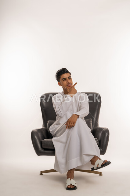 الشعور بالحيرة وعدم التأكد, النظر الى مكان ما بإيماءات التأمل والتمعن والتركيز, بورتريه لشاب مراهق عربي خليجي اماراتي يرتدي الكندورة يجلس على كرسي مريح يضع يده على ذقنه بتعابير التفكير، التخطيط لحل مشكلة ما، خلفية بيضاء