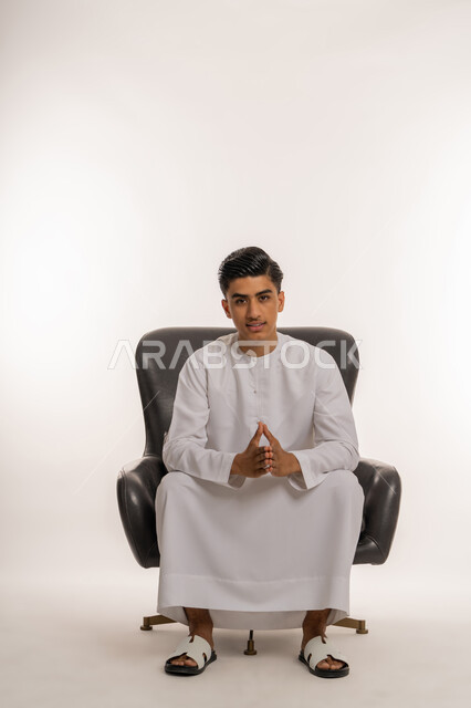 The concept of elegance, masculinity and interest in external appearance, portrait of a smiling young Arab Gulf Emirati teenager wearing a kandura sitting on a comfortable chair with touching hand gestures, looking at the camera with expressions of pleasure and joy, white background