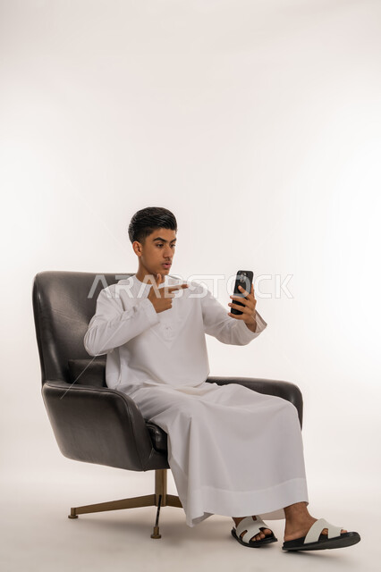 تصفح وسائل التواصل الاجتماعي ومتابعة آخر الأخبار والمستجدات حول العالم، دمج التقنيات الحديثة والتكنولوجيا المتطورة مع الحياة اليومية، بورتريه لشاب مراهق عربي خليجي اماراتي يرتدي الكندورة يجلس على كرسي يشير بإصبع السبابة الى الجوال بتعابير الاندهاش، خلفية بيضاء