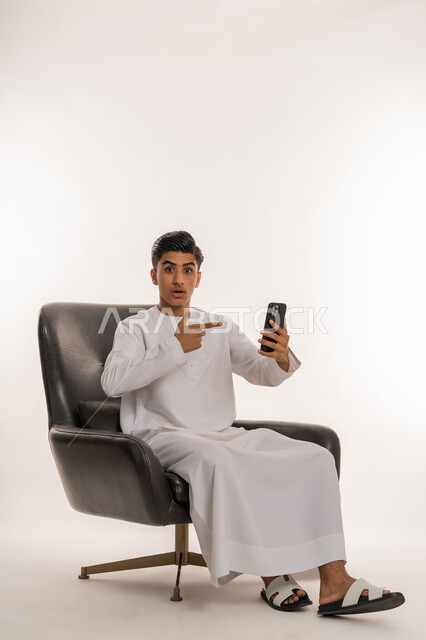 متابعة آخر الأخبار والمستجدات حول العالم، دمج التقنيات الحديثة والتكنولوجيا في الحياة اليومية، بورتريه لشاب مراهق عربي خليجي اماراتي يرتدي الكندورة يجلس على كرسي يشير بإصبع السبابة الى الهاتف المحمول، النظر الى الكاميرا بتعابير الاندهاش والذهول، خلفية بيضاء