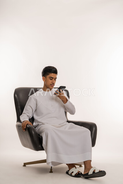 Using technology and modern technical devices, answering calls and communications, portrait of a young Arab Gulf Emirati teenager wearing a kandura sitting on a comfortable chair making a call using a mobile phone, communicating with family and friends via mobile phone, white background
