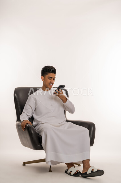Using technology and modern technical devices in daily life, answering calls and communications, portrait of a young Arab Gulf Emirati teenager wearing a kandura sitting on a comfortable chair making a call using a mobile phone, communicating with family and friends via mobile phone, white background