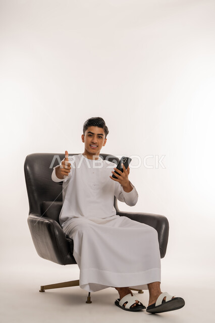 رفع اصبع الابهام للأعلى بتعابير الإعجاب، متابعة آخر الأخبار والمستجدات حول العالم، الاستفادة من التقنيات الحديثة والتكنولوجيا المتطورة، بورتريه لشاب مراهق عربي خليجي اماراتي يرتدي الكندورة يجلس على كرسي يحمل الهاتف المحمول بيده بإيماءات القبول، خلفية بيضاء