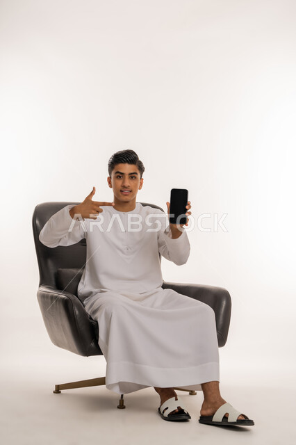 تصفح ومتابعة وسائل التواصل الاجتماعي، دمج التقنيات الحديثة والتكنولوجيا مع الحياة اليومية، بورتريه لشاب مراهق عربي خليجي اماراتي يرتدي الكندورة يجلس على كرسي يشير بإصبع السبابة الى الهاتف المحمول، خلفية بيضاء