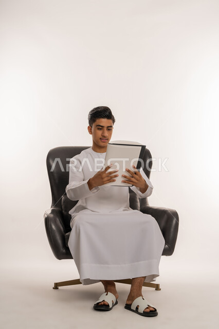 تعابير الاجتهاد والتفوق، تطوير المناهج التعليمية في الامارات العربية المتحدة, بورتريه لشاب مراهق عربي خليجي اماراتي يرتدي الكندورة يجلس على كرسي مريح يقوم بالنظر الى الكتاب ومراجعة الدروس، ايماءات التركيزوالتفكير، خلفية بيضاء
