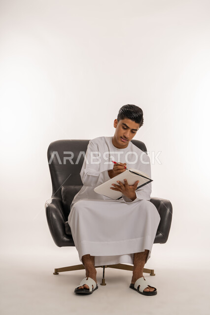 مراجعة الدروس للمذاكرة والاختبارات المدرسية، تطوير المناهج التعليمية في الامارات، تعابير الاجتهاد والتفوق, بورتريه لشاب مراهق عربي خليجي اماراتي يرتدي الكندورة يجلس على كرسي مريح يقوم بالكتابة على الكتاب المدرسي، ايماءات التركيزوالتفكير، خلفية بيضاء