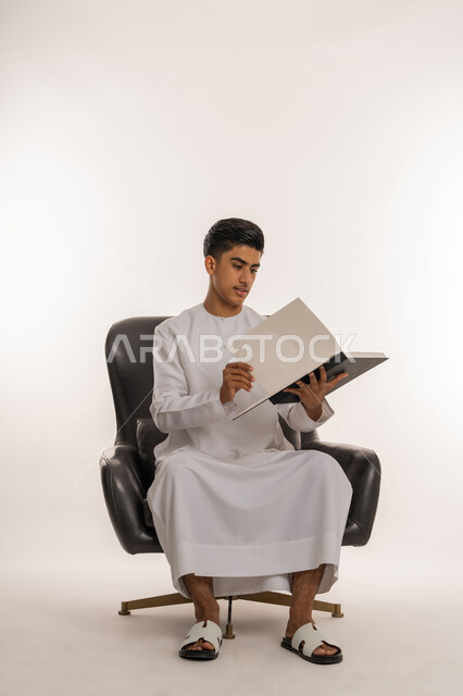 تعابير الاجتهاد والتفوق، تطوير المناهج التعليمية في الامارات العربية المتحدة, بورتريه لشاب مراهق عربي خليجي اماراتي يرتدي الكندورة يجلس على كرسي مريح يقوم بالنظر الى الكتاب ومراجعة الدروس، ايماءات التركيزوالتفكير، خلفية بيضاء