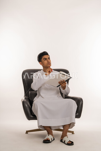 مراجعة الدروس للمذاكرة والاختبارات المدرسية، تطوير المناهج التعليمية في الامارات، إيماءات الاجتهاد والتفوق, بورتريه لشاب مراهق عربي خليجي اماراتي يرتدي الكندورة يجلس على كرسي مريح يحمل بيده الكتاب، النظر الى الاعلى بتعابير التفكير، خلفية بيضاء