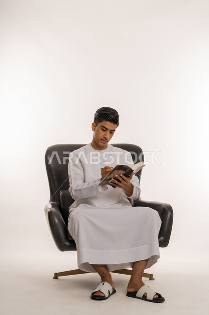 تعابير الاجتهاد والتفوق، مراجعة الدروس للمذاكرة والاختبارات المدرسية، تطوير المناهج التعليمية في الامارات, بورتريه لشاب مراهق عربي خليجي اماراتي يرتدي الكندورة يجلس على كرسي مريح يقوم بالكتابة على الكتاب المدرسي، ايماءات التركيزوالتفكير، خلفية بيضاء