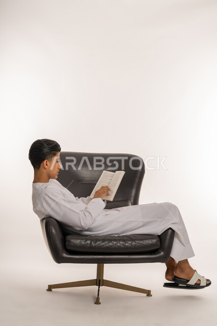 مراجعة الدروس للمذاكرة والاختبارات المدرسية، إيماءات الاجتهاد والتفوق، تطوير المناهج التعليمية في الامارات, بورتريه لشاب مراهق عربي خليجي اماراتي يرتدي الكندورة مستلقي على الكرسي ينظر الى الكتاب المدرسي بتعابير التفكير، خلفية بيضاء