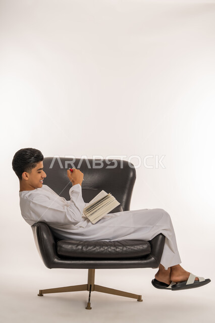 مراجعة الدروس للمذاكرة والاختبارات المدرسية، إيماءات الاجتهاد والتفوق، تطوير المناهج التعليمية في الامارات, بورتريه من الجانب لشاب مراهق عربي خليجي اماراتي يرتدي الكندورة مستلقي على الكرسي ينظر الى الكتاب المدرسي بتعابير التفكير، خلفية بيضاء