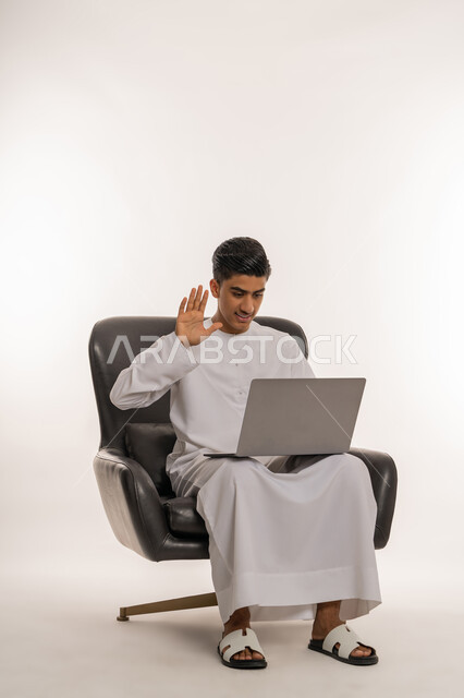 القاء التحية والترحيب والسلام، التواصل مع الاهل والاصدقاء، استخدام التقنيات والاجهزة الحديثة، بورتريه لشاب مراهق عربي خليجي اماراتي يرتدي الكندورة يحمل بيده الابتوب يقوم بإجراء مكالمة فيديو عبر تطبيقات التواصل الاجتماعي، ايماءات الاندماج والسرور، خلفية بيضاء