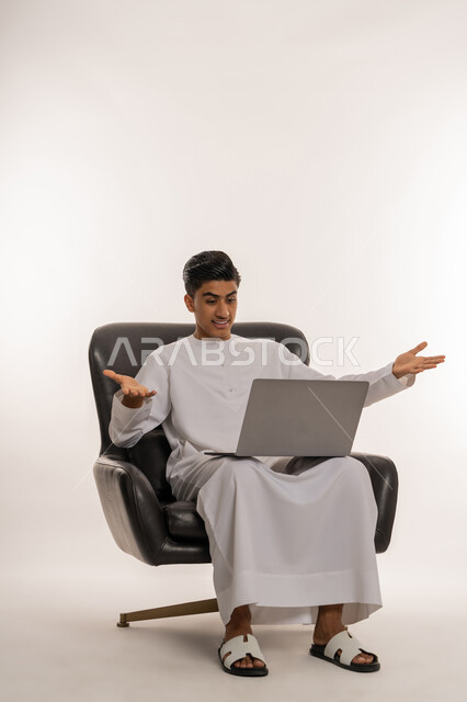The concept of kinship, communicating with family and friends via computer, using modern technologies and devices, portrait of a young Arab Gulf Emirati teenager wearing a kandura making a video call via social media applications, opening his arms with expressions of questioning and surprise, white background