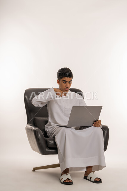 دمج التكنولوجيا في الحياة اليومية، بورتريه لشاب مراهق عربي خليجي اماراتي يرتدي الكندورة يقوم بإجراء مكالمة فيديو عبر تطبيقات التواصل الاجتماعي باستخدام جهاز اللابتوب، الإشارة بإصبع السبابة على الحاسوب المحمول، إيماءات الاندهاش والذهول، خلفية بيضاء