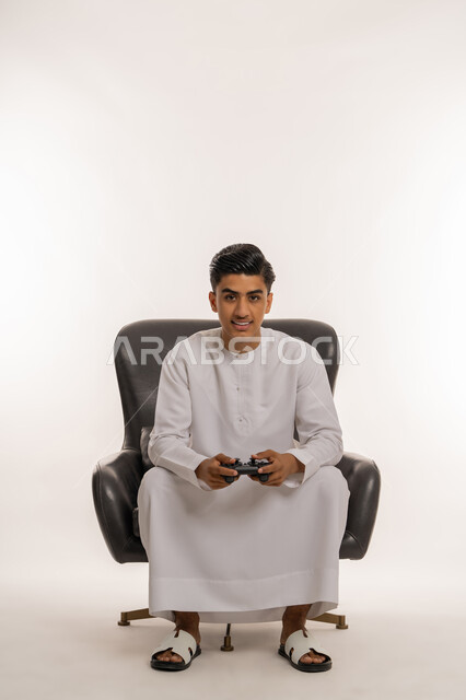 قضاء أوقات ممتعة في ألعاب الفيديو الترفيهيه، بورتريه لشاب مراهق عربي خليجي اماراتي مبتسم يرتدي الكندورة البيضاء يحمل بيده عصا التحكم بإيماءات الفرح والاستمتاع والاندماج مع الاجهزة الحديثة، الجلوس على كرسي مريح، خلفية بيضاء