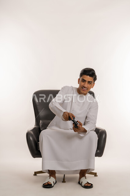 دمج التسلية بالتكنولوجيا المتطورة، قضاء أوقات ممتعة في ألعاب الفيديو الترفيهيه، بورتريه لشاب مراهق عربي خليجي اماراتي مبتسم يرتدي الكندورة البيضاء يحمل بيده عصا التحكم بإيماءات الفرح والاستمتاع والاندماج مع الاجهزة الحديثة، الجلوس على كرسي مريح، خلفية بيضاء