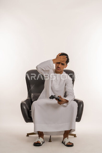 تقنيات إلكترونية ترفيهيه، بورتريه لشاب مراهق عربي خليجي اماراتي يرتدي الكندورة البيضاء يحمل بيده عصا التحكم يضع يده على رأسه بتعابير الخسارة والهزيمة، تجربة ممتعة في الأجهزة الحديثة، ايماءات الاستمتاع والاندماج والتفاعل مع ألعاب الفيديو، خلفية بيضاء