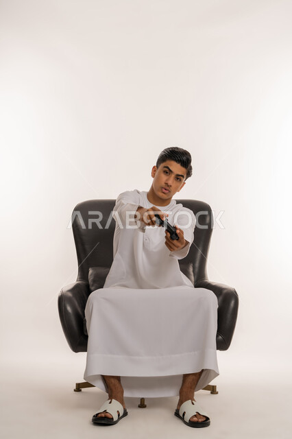 قضاء وقت ممتع  في الأجهزة الحديثة، بورتريه لشاب مراهق عربي خليجي اماراتي يرتدي الكندورة البيضاء يحمل بيده عصا التحكم ينظر للكاميرا بتعابيرالعبوس  والانزعاج، ايماءات الاندماج والتفاعل مع ألعاب الفيديو الترفيهية، خلفية بيضاء
