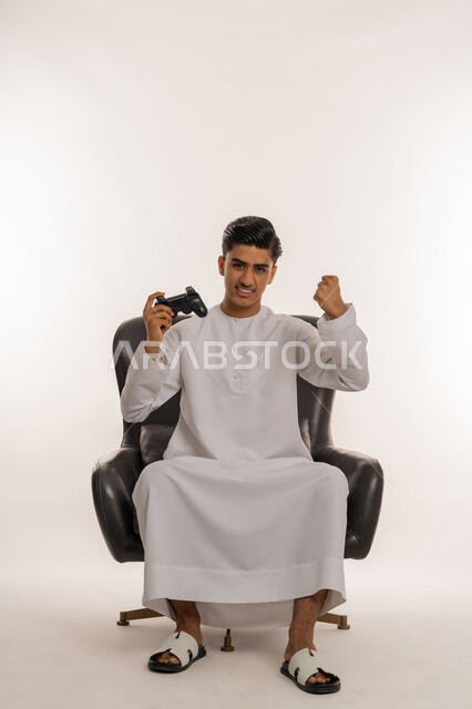 Raising a fist with expressions of victory and triumph, entertainment electronic technologies, portrait of a young Arab Gulf Emirati teenager wearing a white kandura sitting on a comfortable chair holding a joystick in his hand with gestures of enjoyment and integration with video games, spending enjoyable times on modern devices, white background