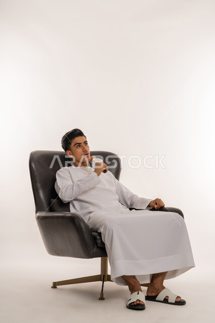 Recording a singing voice clip, gestures of integration with singing, enjoying the performance of vocal talents, portrait of a young Emirati Arab Gulf teenager wearing a kandura holding a microphone in his hand and singing enthusiastically, practicing the talent of singing, sitting on a comfortable chair, white background