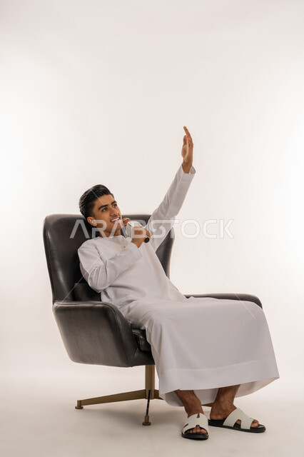 ايماءات الاندماج والتفاعل والرقص مع الغناء، الاستمتاع بأداء المواهب الصوتية، تسجيل مقطع صوتي غنائي، بورتريه لشاب مراهق عربي اماراتي خليجي يرتدي الكندورة يمسك بيده المايكروفون و يغني بحماس، ممارسة موهبة الطرب، الجلوس على كرسي مريح، خلفية بيضاء