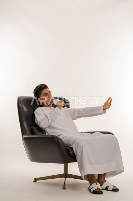 Gestures of integration, interaction and dancing with singing, enjoying the performance of vocal talents, recording a singing audio clip, portrait of a young Emirati Gulf Arab teenager wearing a kandura holding a microphone in his hand and singing enthusiastically, practicing the talent of singing, sitting on a comfortable chair, white background
