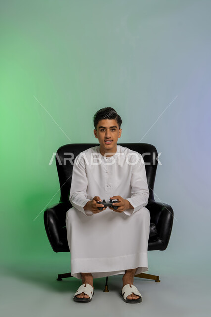 قضاء أوقات ممتعة في ألعاب الفيديو الترفيهية، بورتريه لشاب مراهق عربي خليجي اماراتي مبتسم يرتدي الكندورة البيضاء يحمل بيده عصا التحكم بإيماءات الفرح والاستمتاع والاندماج مع الاجهزة الحديثة، الجلوس على كرسي مريح، خلفية ملونة