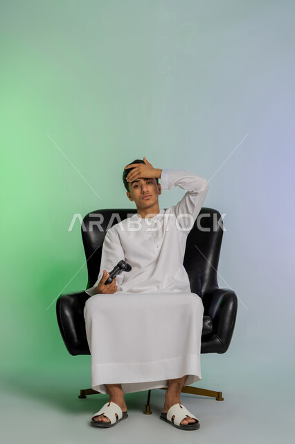 تقنيات إلكترونية ترفيهيه، بورتريه لشاب مراهق عربي خليجي اماراتي يرتدي الكندورة البيضاء يحمل بيده عصا التحكم يضع يده على رأسه بتعابير الخسارة والهزيمة، تجربة ممتعة في الأجهزة الحديثة، ايماءات الاستمتاع والاندماج والتفاعل مع ألعاب الفيديو، خلفية ملونة