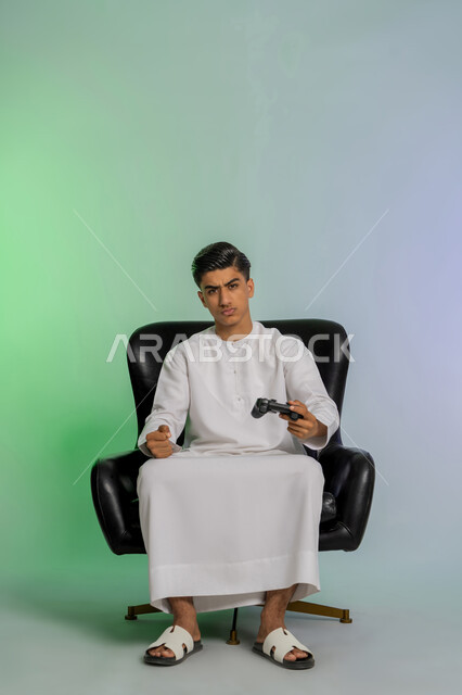 تجربة ممتعة ومثيرة في الأجهزة الحديثة، تقنيات إلكترونية ترفيهيه، بورتريه لشاب عربي خليجي اماراتي يرتدي الكندورة البيضاء يحمل بيده عصا التحكم ينظر للكاميرا بتعابير العبوس والانزعاج، ايماءات الاندماج والتفاعل مع ألعاب الفيديو، خلفية ملونة