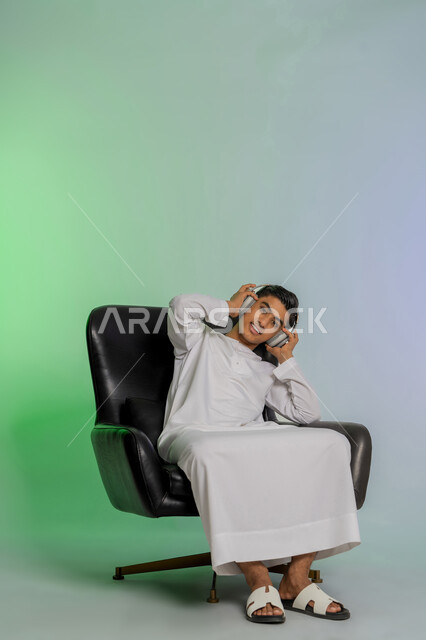 إيماءات  الاندماج والطرب، مفهوم الإصغاء والاستمتاع بإستخدام الهيدفون، قضاء أوقات ممتعة بالإستماع للمواهب الغنائية، بورتريه لشاب مراهق عربي خليجي اماراتي يرتدي الكندورة البيضاء يضع سماعة الرأس، الجلوس على كرسي مريح، خلفية ملونة