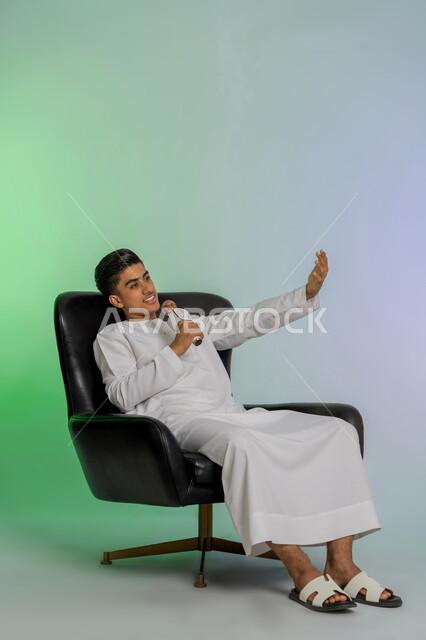 تسجيل مقطع صوتي غنائي، ايماءات الاندماج والتفاعل مع الغناء،  الاستمتاع بأداء المواهب الصوتية، بورتريه لشاب مراهق عربي اماراتي خليجي يرتدي الكندورة يمسك بيده المايكروفون و يغني بحماس، ممارسة موهبة الطرب، الجلوس على كرسي مريح، خلفية ملونة
