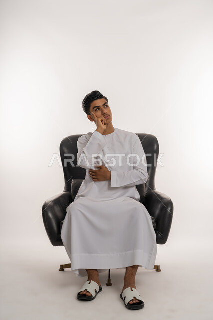 Trying to find a solution to a problem, contemplating, concentrating and planning for the future, portrait of a young Arab Gulf Emirati teenager wearing a kandura sitting on a comfortable chair and putting his hand on his cheek with expressions of thinking and contemplation, mind wandering somewhere, looking away to remember something, gestures of confusion and anxiety, white background