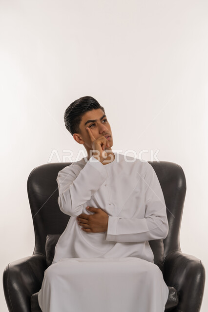 التمعن والتركيز والتخطيط للمستقبل، بورتريه مقرب لشاب مراهق عربي خليجي إماراتي يرتدي الكندورة يجلس على كرسي مريح ويضع يده على خده بتعابير التفكير والتأمل، شرود الذهن إلى مكان ما, النظر بعيداً لتذكر شيء، إيماءات الحيرة والقلق، محاولة إيجاد حل مشكلة، خلفية بيضاء